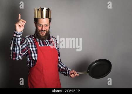 King of cooking with finger up, funny bearded man in paper crown and cook apron with empty frying pan is ready to tech a lesson Stock Photo