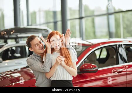 Emotional shot of loving successful husband making surprise for his wife, buying her a new car at dealership. Surprised red haired young woman screami Stock Photo