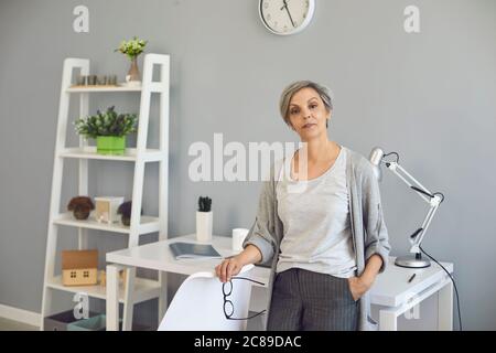 Senior woman in casual clothes looks at the camera on the background of the workplace at home office. Stock Photo