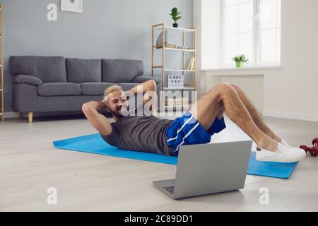 Young guy working out to online video tutorial on laptop at home. Sporty man doing abs exercises on yoga mat indoors Stock Photo