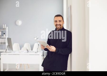 A positive guy smiling standing with a tablet in his hand in the office at the workplace. Stock Photo