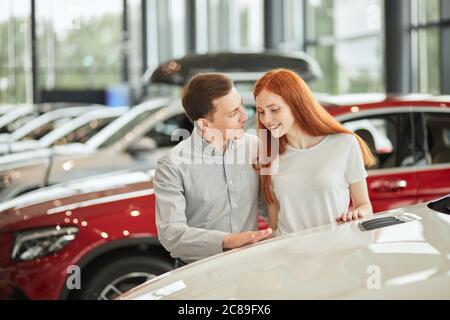 Happy caucasian couple chooses their first family car to buy in a showroom. Successful businessman asking his beautiful red-haired wife whether she li Stock Photo