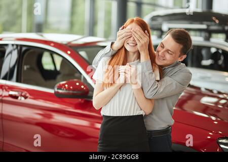 Caucasian charming couple stand in front of new red car, man embracing his wife covered her eyes with his hands Stock Photo