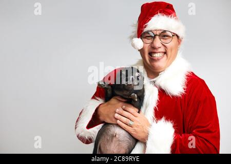 Happy Asian man in spectacles and Santa Claus suit holds black little piggy in hands smiling broadly at camera wishing Merry christmas, isolated over Stock Photo