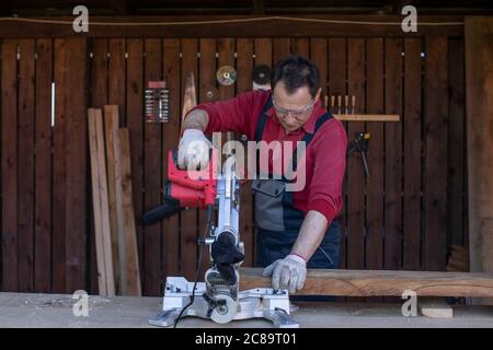 Adult man in protective overalls and glasses cuts wooden beam using circular saw Stock Photo