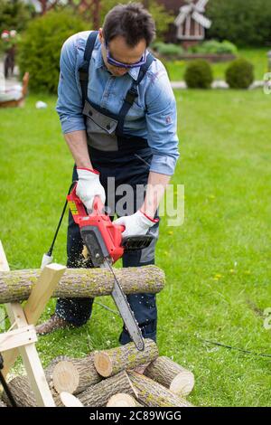 Middle-aged man in overalls cuts an aspen trunk into pieces with a chainsaw. Stock Photo