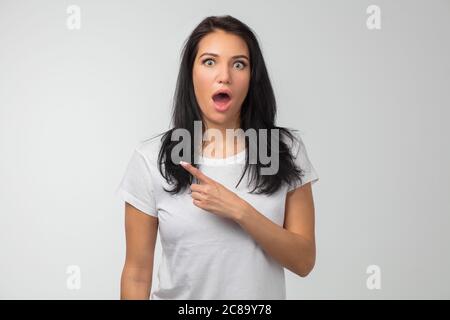 Amazed female with long black hair wearing casual clothes, looking with bugged eyes and widely opened mouth, pointing withat copy space. close up phot Stock Photo