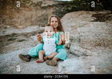 happy young girl sitting and holding baby brother on a large rock Stock Photo