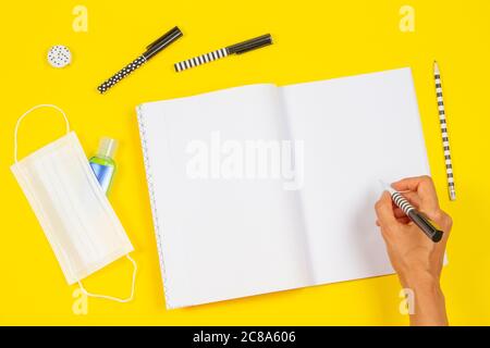 Top view to woman hand writing on open notebook. School supplies, hand sanitizer and medical face mask on yellow background Stock Photo