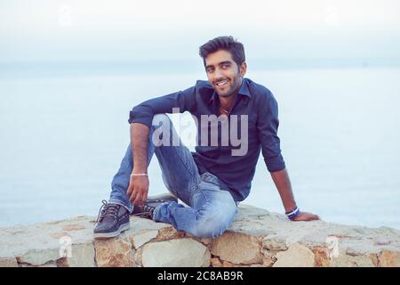 Man smiling looking to the side happy celebrating freedom while sitting on a concrete wall bridge above the sea. Positive person, peace mind concept. Stock Photo