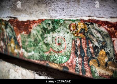 Teotihuacan Murals San Juan Teotihuacan Mexico | SAN JUAN TEOTIHUACÁN, Mexico — A shadowed close-up of a Teotihuacan mural fragment reveals a bird- or serpent-headed figure painted in red, ochre, and green on weathered lime plaster. Murals of this kind decorated residential apartment compounds throughout the city, reflecting a shared artistic vocabulary of deities and ritual imagery. Teotihuacan's mural tradition is among the most extensive to survive from ancient Mesoamerica. Stock Photo