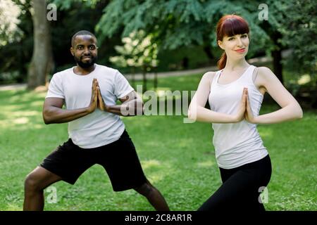 Young multiethnic couple, African man and Caucasian woman, doing yoga in nature outdoors, standing with hands together in prayer position. Healthy Stock Photo