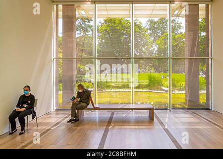 The Fondation Beyeler's spacious park in Riehen, Switzerland, with its pond and lawn, is visible through the floor-to-ceiling glass façade of the exhibition building. Two visitors wearing face masks sit on a wooden bench in the foreground, one leafing through a companion booklet to the special exhibition "Edward Hopper". The museum building, designed by Renzo Piano, opened in 1997. Stock Photo