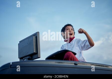 Excited Schoolboy fist pumps while working on a homework using computer on top of car roof, New Normal Education concept Stock Photo