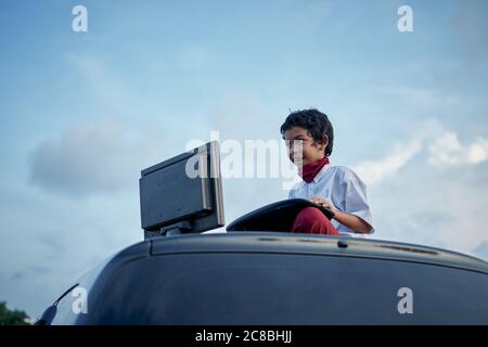 Boy in school uniform playing internet games on top of car roof, New Normal Education concept Stock Photo