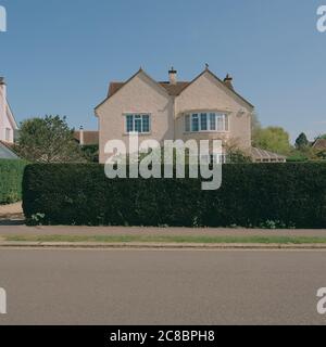 A typical M Shaped / Double Pitched / Double Gabled Roof suburban house half hidden behind a green garden hedge and residential street with blue sky Stock Photo