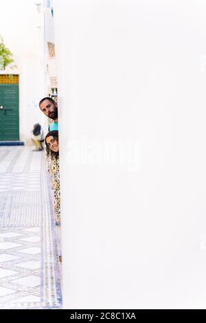 Vertical photo of two tourists sticking their heads out from behind a white wall of a building in Morocco Stock Photo