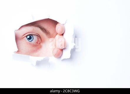 Mans blue eye peering through through ripped paper Stock Photo
