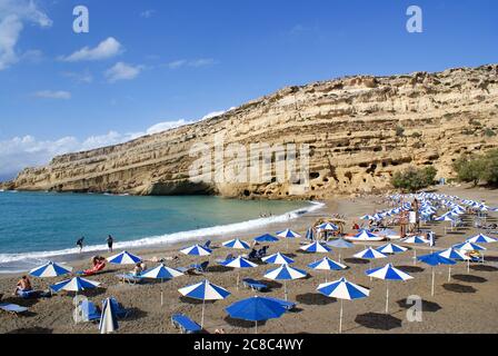Matala, Crete / Greece / October 15 2010 : Tourists relax in the shade of colorful blue and white parasols on the beach at Matala, Crete.  Late summer Stock Photo