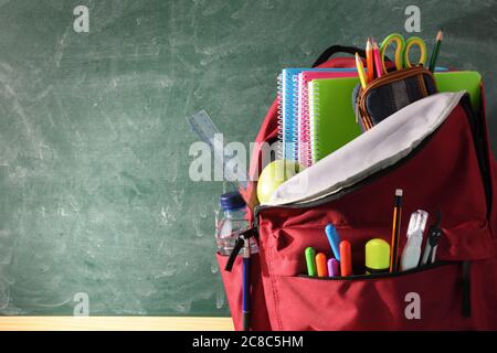 Backpack full of school supplies on wood table and green blackboard with blank space for writing front view Stock Photo