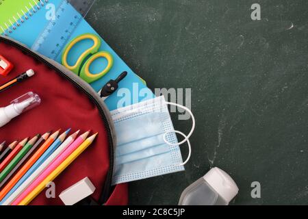 Backpack full of school supplies on green blackboard with protection mask and hydroalcoholic gel detail. Top view Stock Photo