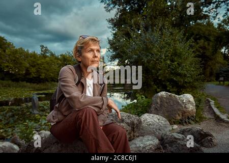 An elderly woman sits on the rocks by the pond and watches the sky after a summer storm. Stock Photo