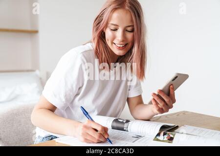 Image of smiling caucasian woman using smartphone while doing homework with exercise books in home Stock Photo