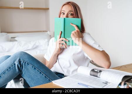 Image of shocked pretty woman with pink hair reading book while doing homework at home Stock Photo
