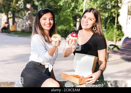 two attractive and cheerful girls enjoying delicious donuts in city park in a positive and energetic mood Stock Photo
