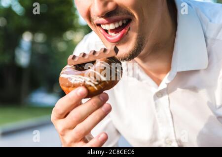 close up view of handsome caucasian young man in white shirt enjoying delicious chocolate donut in city park. junk but tasty food for positive mood Stock Photo