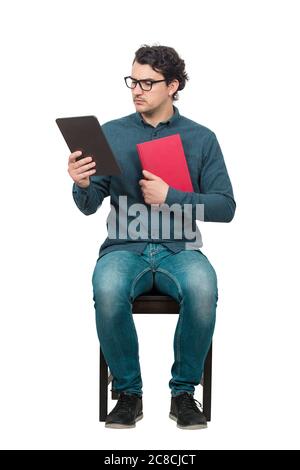 Curious man student or teacher, seated on a chair using a PC tablet while holds a red book. Choosing between a traditional textbook and a modern Ebook Stock Photo