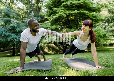 Two smiling sporty friends, multiethnic couple, African man and Caucasian woman, giving high five to each other, while doing push up or plank on Stock Photo