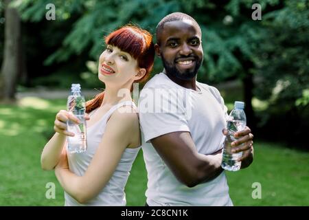 Sport, fitness and family concept. Close up of cheerful smiling multiethnical couple with bottles of water, posing to camera during their break after Stock Photo