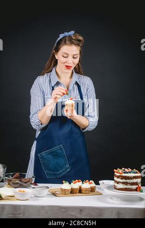 Happy young woman in denim apron decorating delicious cake with berries on black background in studio Stock Photo