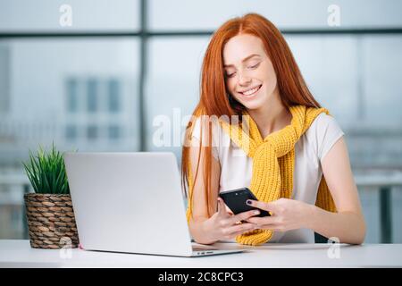 Young business woman sitting in office at desk, looking at graphics and uses smartphone. On table is laptop and tablet computer. Teamwork. Stock Photo