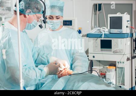 Surgeon performing cosmetic surgery on nose in hospital operating room. Surgeon in mask wearing surgical loupes during medical procedure. nose augment Stock Photo