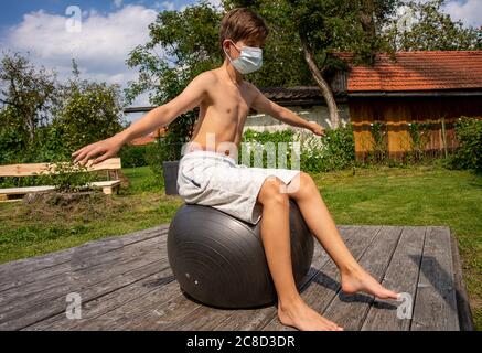 A boy wearing a facemask balances on a ball while doing back excercises Stock Photo