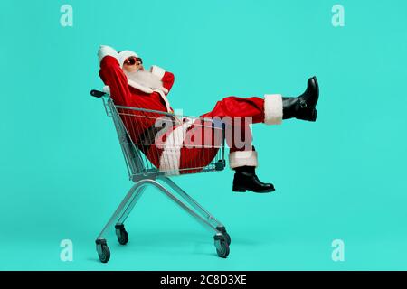 Side view of funny Santa Claus in black sunglasses and costume making faces. Old man having fun, sitting and relaxing in shopping cart on blue studio background. Concept of holidays, fun. Stock Photo