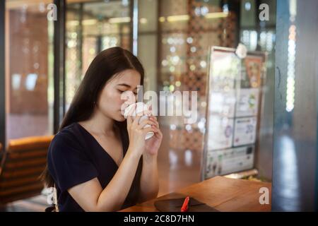 Long hair woman in dark blue dress sitting in the cafe waiting for her boyfriend, drinking hot coffee, drink coffee with absent-minded. Stock Photo