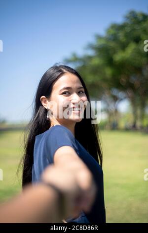 Long hair woman in dark blue dress lively walking on the grass field in the park and holding her boyfriend's hand then smile. Stock Photo