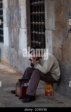 Damascus, Syria 03/28/2010: A poor young Syrian man in torn clothes is sitting on an empty can  in front of his shoe shine stand. He is taking a bite Stock Photo