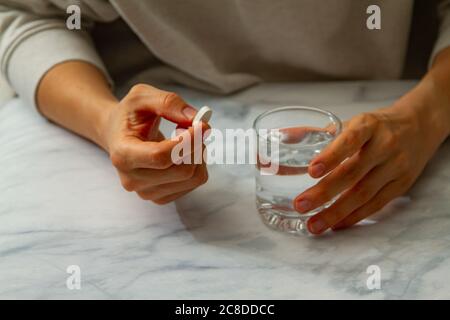 A woman is seen as she is holding a pill in one hand and a glass of water on the other hand. She is about to take the medication with a sip of water.