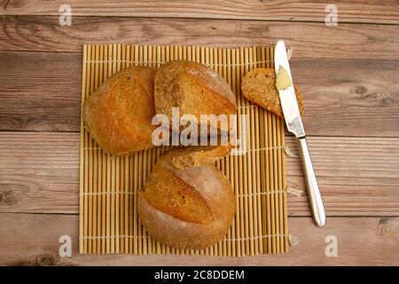 close up image of home made whole wheat bread loaves fresh out of oven. They are cooling on a bamboo mat on wooden table. A butter knife with butter. Stock Photo