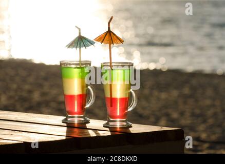 Glasses with multi-colored cocktails on a wooden table in the sun, the beach and the sea in the background Stock Photo