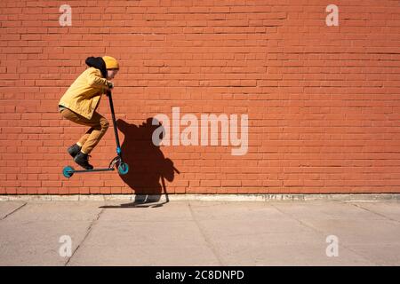 Boy performing stunt with push scooter on sidewalk against brick wall during sunny day Stock Photo