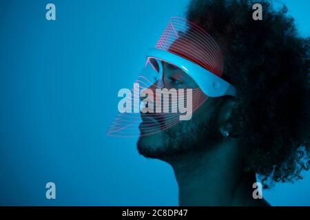 Thoughtful young man wearing red led mask against wall at home Stock Photo