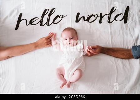 Happy cute baby girl holding hands of parents while lying on blanket under Hello World text at home Stock Photo