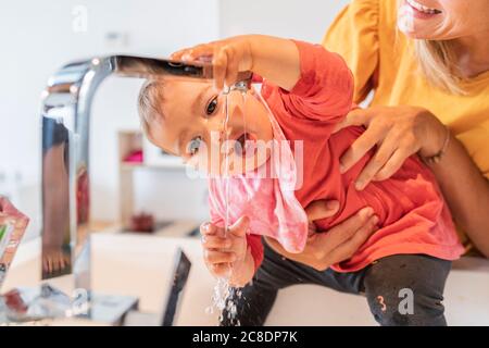 Close-up of mother holding cute baby girl drinking water through faucet in kitchen sink Stock Photo