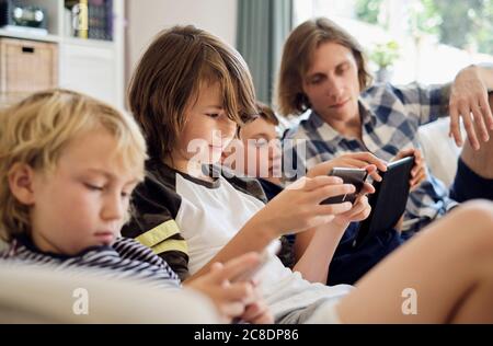 Father looking at boys playing games on smart phones and digital tablet in living room Stock Photo