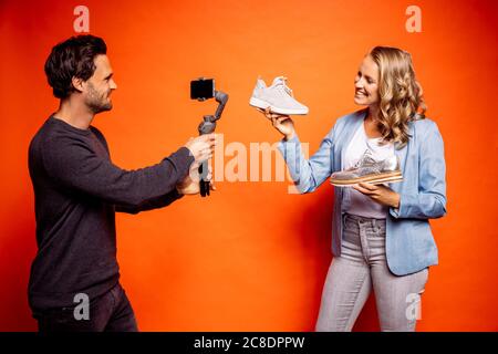 Smiling woman showing shoes while man photographing her through smart phone against orange background Stock Photo